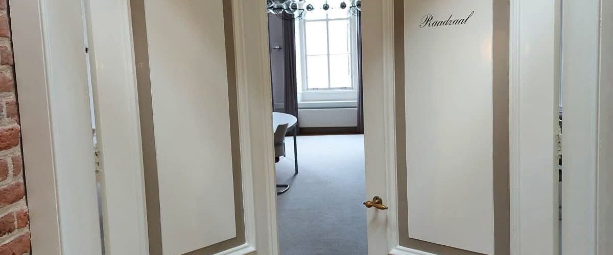 Twee witte deuren die leiden naar de raadzaal van Hattem staan open op een kier. Door deze kier zie je een raam, een kandelaar en een stukje van een vergadertafel.