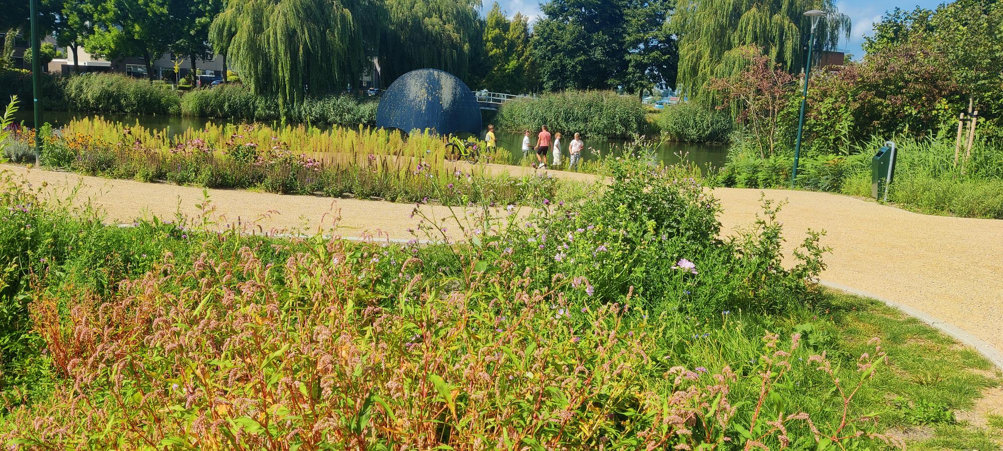Je ziet een groen park met diverse soorten bloemen en grassen, die bloeien naast een kiezelpad. In de achtergrond wandelt iemand, terwijl twee kinderen in een vijver staan te vissen en hun ouders toekijken. Achter de vijver staan enkele rietkluiten en wilgen.